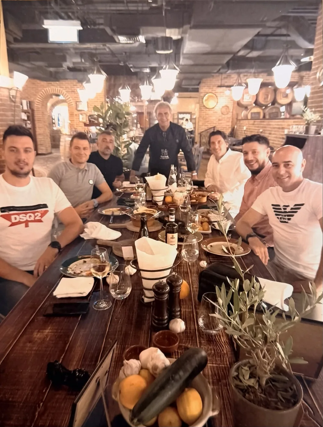 Una tavolata conviviale: lo chef in piedi al capotavola, ospiti seduti attorno a un grande tavolo di legno.