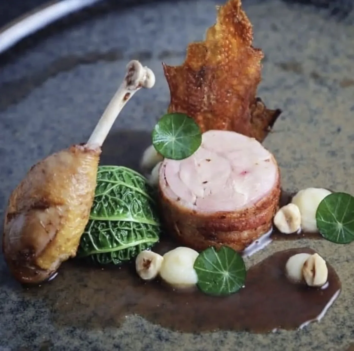 Anatra in due cotture con cavolo verza, nocciole e salsa al fondo, su piatto scuro.